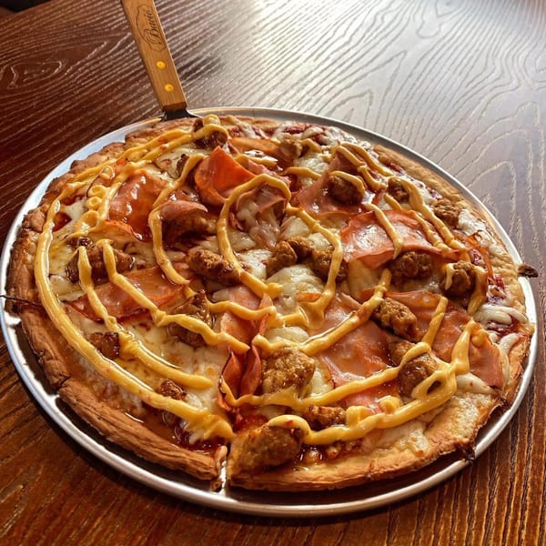 A whole pizza with ham, sausage, and a mustard drizzle on a metal pan with a pizza server.